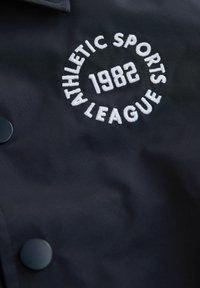 Chaqueta azul marino con un logo circular bordado en blanco que dice "ATHLETIC SPORTS LEAGUE 1982" con botones a presión negros para cierre.