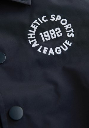 Marinblå jacka med en cirkulär vit broderad logotyp som lyder "ATHLETIC SPORTS LEAGUE 1982" och svarta tryckknappar för stängning.