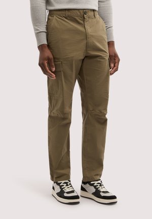 Hombre vistiendo pantalones cargo verde oliva y zapatillas blancas y negras de pie contra un fondo liso.