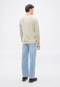 Világos bézs kötött pulóver texturált ujakkal, világoskék, bőszárú farmerral és sötétbarna cipővel. Hátulnézetből.