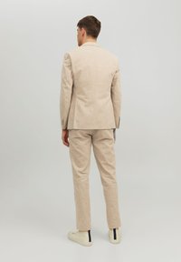 Completo beige chiaro con una finitura texturizzata, caratterizzato da una giacca monopetto e pantaloni affusolati. Indossato con sneakers bianco sporco.