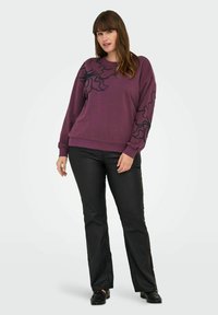 Sudadera morada con diseño de líneas abstractas negras en las mangas, con cuello redondo y puños acanalados, combinada con pantalones acampanados negros y zapatos.