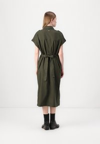 Rochie din in de culoare verde măslin, cu mâneci scurte, cordon la talie și tiv până la gambe. Purtați cu botine negre la gleznă, cu toc masiv.