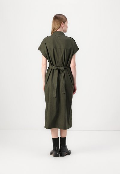 Robe en lin vert olive avec des manches courtes, ceinture à nouer à la taille et ourlet au niveau des mollets. Portée avec des bottines noires à talons épais.