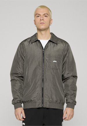 Jeune personne aux cheveux blonds courts portant une veste zippée gris foncé sur une chemise blanche et un pantalon noir, regardant vers l'avant avec une expression neutre.