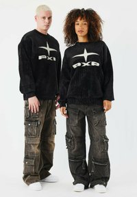 Schwarzer Fleece-Hoodie mit weißem "PXP"-Logo, kombiniert mit dunklen Cargohosen mit mehreren Taschen und Reißverschlüssen, getragen mit weißen Sneakers.
