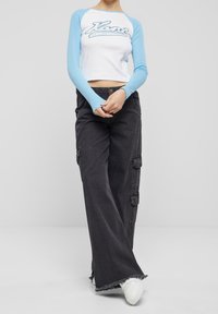 Valkoinen lyhyt cropped-toppi, jossa on siniset pitkät hihat ja "Levi's"-logo. Mustat leveälahkeiset cargo-housut, joissa on taskuja ja hapsutetut lahkeet. Valkoiset lenkkarit.