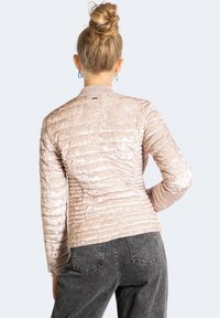 Žena s blond vlasy v drdolu, oblečená v lehce růžové prošívané bundě a tmavě šedých džínech, stojí s jednou rukou na bocích, s zády k fotografii.