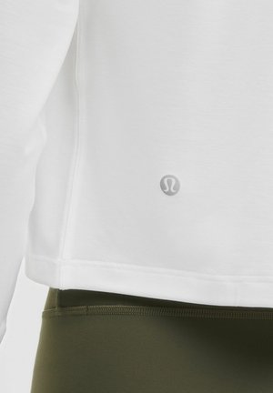 Baltas, minkštas audinys ilgomis rankovėmis marškinėliai su apvalia apačia ir subtiliu logotipu; derinami su tamsiai alyvuogių žalios spalvos, tampriais leggingais su siuvinėjimo detalėmis.