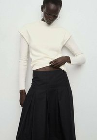 Haut à manches longues crème avec des accents aux épaules, associé à une jupe noire plissée. Les textures sont douces et le contraste des couleurs est marqué.