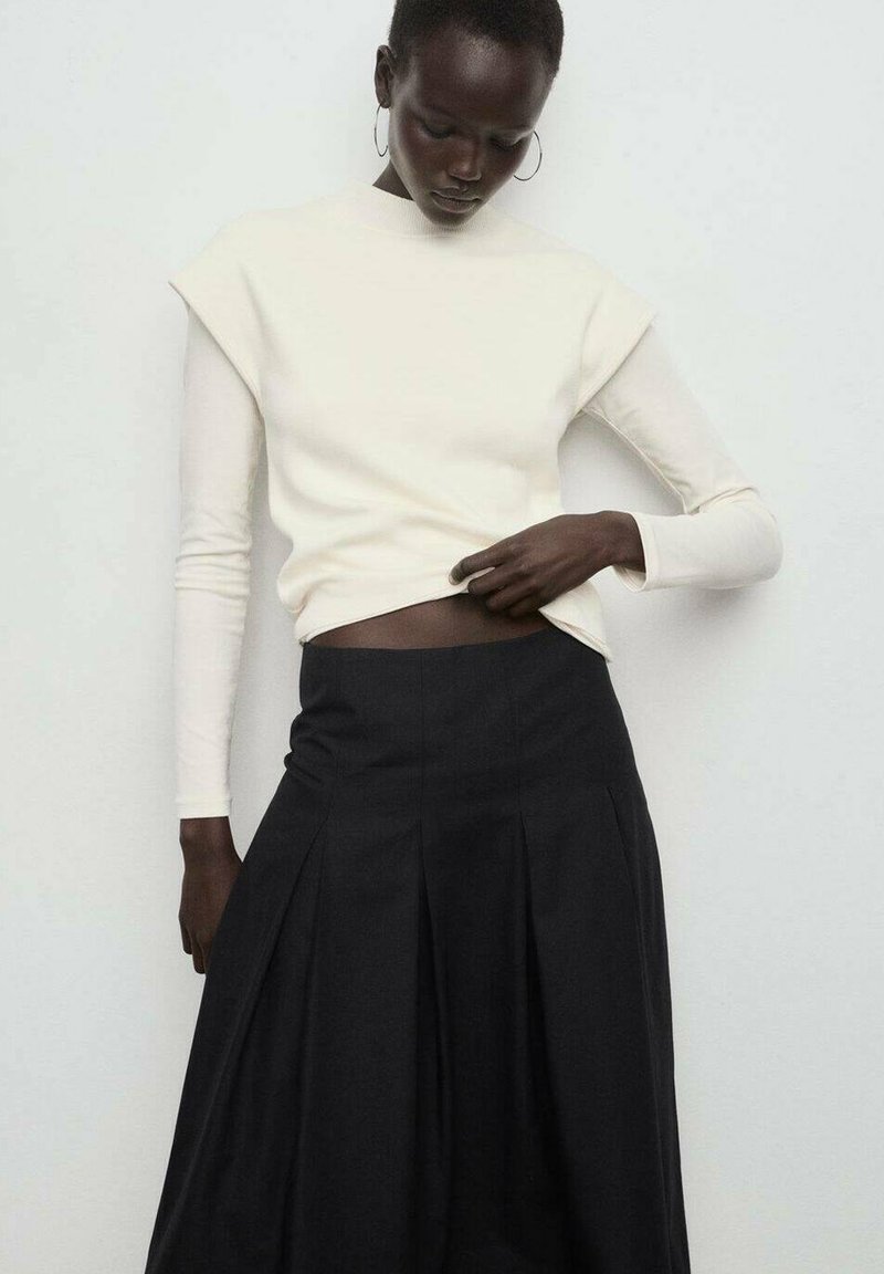 Haut à manches longues crème avec des accents aux épaules, associé à une jupe noire plissée. Les textures sont douces et le contraste des couleurs est marqué.
