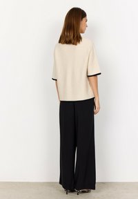 Kvinna med axellångt brunt hår bär en beige kortärmad topp och svarta vida byxor, står mot en vit vägg.