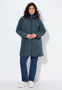 Donna che indossa un cappotto imbottito scuro con zip, un dolcevita blu, jeans blu dritti e sneakers bianche, in piedi contro uno sfondo bianco semplice.