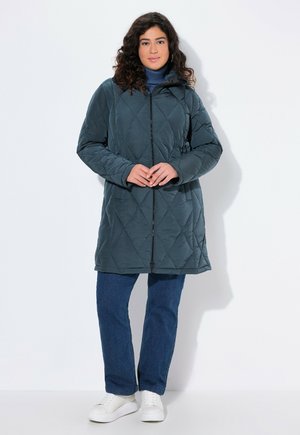 Mujer vestida con un abrigo acolchado oscuro con cremallera, un suéter de cuello alto azul, jeans rectos azules y zapatillas blancas, de pie frente a un fondo blanco liso.
