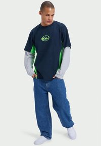 T-shirt azul marinho e verde com um logótipo, sobreposto a uma camisa de manga longa cinza claro, combinado com umas calças de ganga azuis largas e meias brancas.