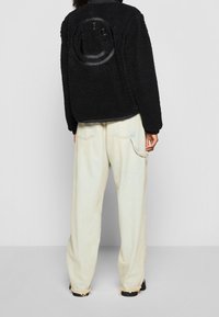 Person som bär lösa, ljusa denimbyxor och en svart fleecejacka med en glansig smiley face-design på ryggen, står mot en enfärgad bakgrund.