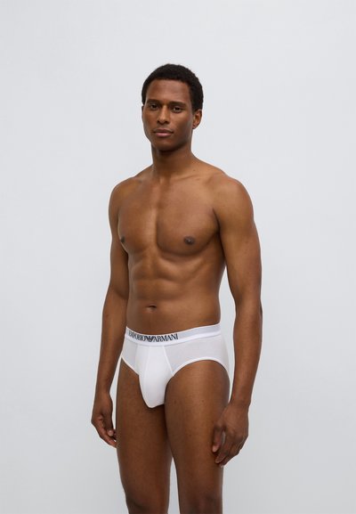 Λευκά βαμβακερά εσώρουχα με μαύρη ταινία μέσης με το λογότυπο Emporio Armani, διαθέτοντας εφαρμοστή σχεδίαση και λεία υφή. Το μοντέλο στέκεται με ουδέτερη έκφραση.
