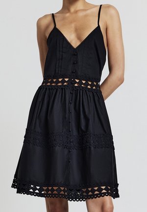 Frau trägt ein ärmelloses schwarzes Kleid mit Knöpfen, Spitzen-Einsätzen an Taille und Saum sowie einem ausgestellten Rock.