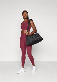 Athletic outfit in burgundy, featuring a cropped tank and high-waisted leggings. Black duffel bag with curved accents and white details. White sneakers.