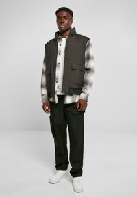 Männeroutfit mit einem dunkelgrünen ärmellosen Weste über einem karierten Flanellhemd, kombiniert mit dunklen Hosen und weißen Sneakers.