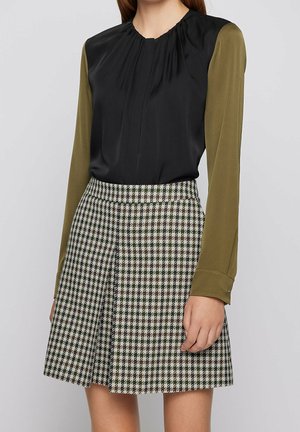 Frau trägt schwarze Bluse mit olivgrünen langen Ärmeln, die in einen hoch taillierten schwarz-weißen Hahnentrittrock gesteckt ist, steht vor einem schlichten Hintergrund.