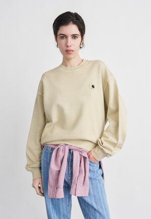 Femeie cu păr scurt și închis la culoare, purtând un hanorac bej, blugi albaștri și o cămașă roz legată în jurul taliei, pe un fundal simplu alb.