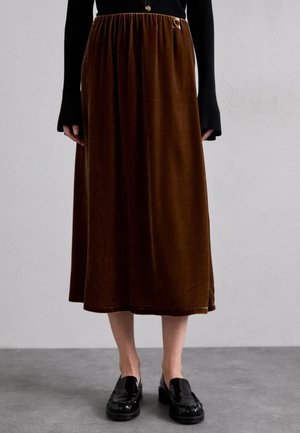Personne portant une jupe midi en velours marron, un haut noir à manches longues côtelé, et des mocassins noirs brillants, debout sur un sol gris.