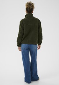Fleece jacket in donker groen, met een hoge kraag en een gestructureerd oppervlak. In combinatie met lichtblauwe wijde jeans, wordt het model van achteren bekeken.
