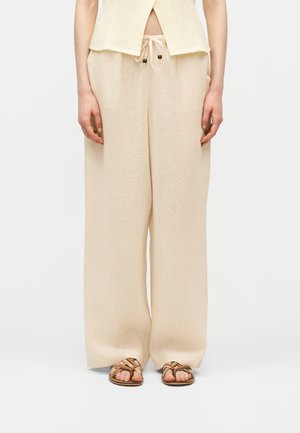 Femme portant un pantalon large en lin beige avec taille à cordon, un haut sans manches jaune clair et des sandales en cuir marron, debout sur un sol blanc.