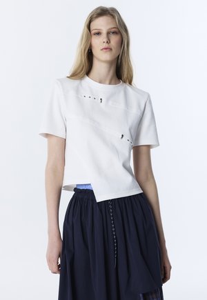 Giovane donna che indossa una maglietta asimmetrica a maniche corte bianca con occhielli metallici e gonna blu navy con vita a coulisse su sfondo bianco.