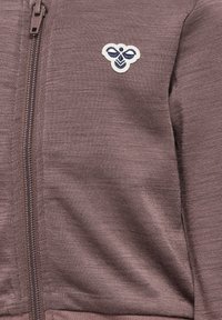 Brun zip-up hoodie gjord av texturerad tyg, med en liten broderad logotyp med ett bi-motiv på vänster bröstområde.