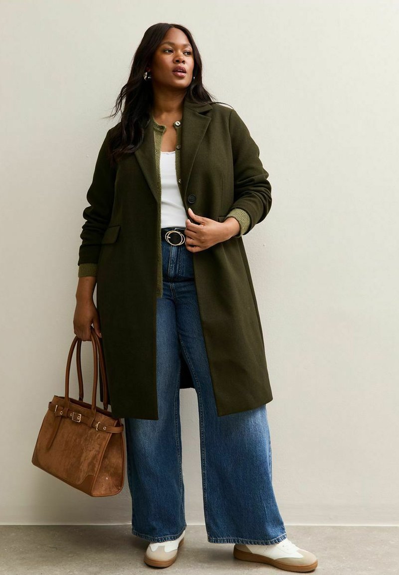 Cappotto di lana verde scuro con bottoni, taglio oversize e polsini a costine. Abbinato a jeans a gamba larga a vita alta e una borsa color beige. Sneakers bianche.