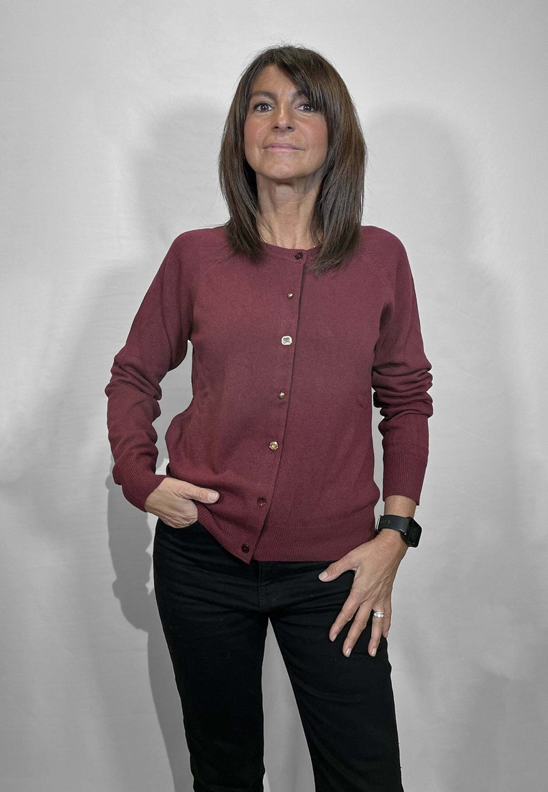 Cardigan color mogano, con trama a coste, sei bottoni sul davanti, scollo arrotondato, abbinato a pantaloni neri, in piedi davanti a uno sfondo chiaro.