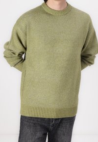 Sweter z dekoltem typu crew neck w miękkim oliwkowym kolorze, dziany, z prążkowanymi mankietami i obrębem. Noszony na ciemnoszarych dżinsach, charakteryzujący się luźnym krojem i teksturowaną tkaniną.