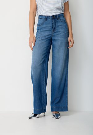 Woman wearing high-waisted, wide-leg blue jeans, striped short-sleeve shirt, and silver pointed-toe heels standing against white background.