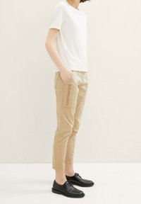White short-sleeve top, beige cropped trousers with side zip pockets, and black lace-up shoes. Smooth fabric and clean lines throughout.