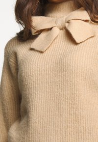 Beige Pullover mit geripptem Muster, hohem Kragen und einer Schleifenapplikation am Kragen. Hergestellt aus weichem, warmem Material.