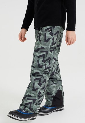 Personne portant un pantalon de neige vert et noir à motif géométrique et des bottes d'hiver noires avec des accents bleus, debout sur un fond blanc uni.
