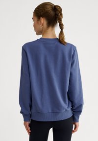 Felpa blu con vestibilità rilassata, polsini e orlo a coste, e texture liscia. Presenta un collo rotondo e un design semplice, senza grafiche.