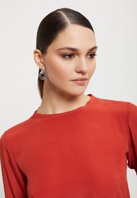 Crvena bluzica od glatkog, laganog materijala s okruglim izrezom. Model nosi kružne metalne naušnice koje prikazuju suvremen dizajn.