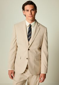 Traje beige a medida en tejido ligero con cierre de dos botones, dos bolsillos y solapas con muesca. Combinado con una camisa blanca y una corbata a rayas.