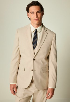 Traje beige a medida en tejido ligero con cierre de dos botones, dos bolsillos y solapas con muesca. Combinado con una camisa blanca y una corbata a rayas.