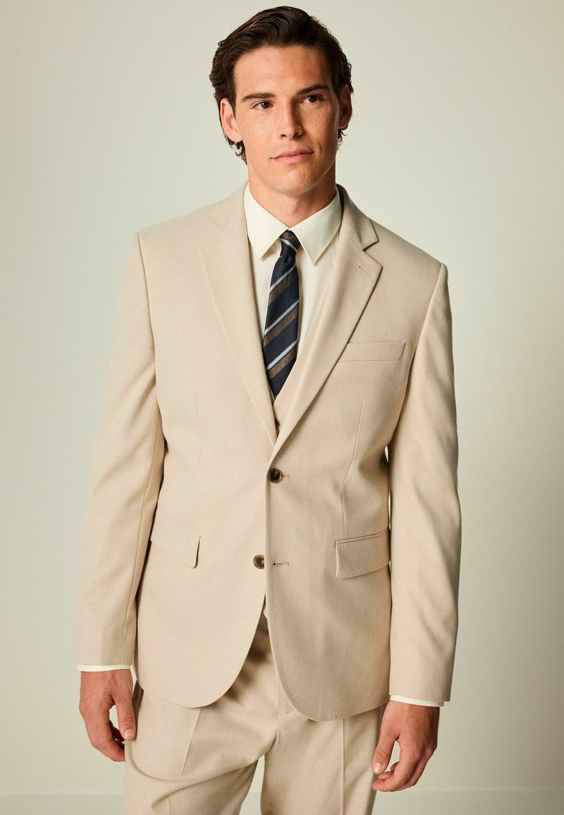 Traje beige a medida en tejido ligero con cierre de dos botones, dos bolsillos y solapas con muesca. Combinado con una camisa blanca y una corbata a rayas.