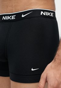 Κοντινό πλάνο ατόμου που φοράει μαύρα boxer briefs Nike με λογότυπο στη μέση και στο πόδι, δείχνοντας μέρος της κοιλιάς και του χεριού.