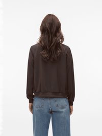 Femme aux longs cheveux bruns ondulés portant un haut noir à manches longues et un jean bleu, debout de dos sur un fond blanc uni.