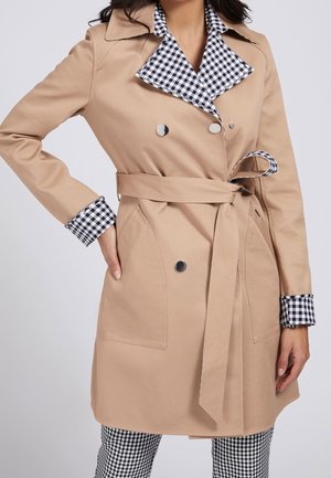 Gabardina beige con cinturón, con cuello y puños de cuadros vichy en blanco y negro, llevada sobre pantalones a juego de cuadros vichy.