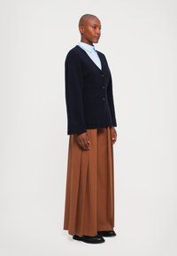 Kvinna med rakad skalle bär en marinblå kofta över en ljusblå skjorta, bruna vida plisserade byxor och svarta skor, stående på sidan.