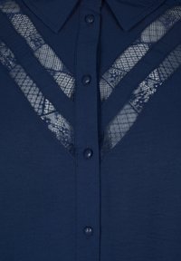 Blouse marine avec des détails en dentelle en motif en V. Dotée d'une fermeture à boutons et d'un col. Texture lisse et tissu léger.