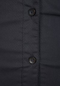 Schwarzes Shirt mit Knopfleiste und zwei runden schwarzen Knöpfen aus einem glatten, strukturierten Stoff, mit sichtbaren Nähten entlang der Knopfleiste.
