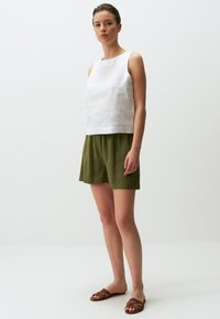 Top senza maniche in lino bianco abbinato a pantaloni corti verde oliva, con una vestibilità rilassata. Il modello indossa sandali marroni con cinturini su uno sfondo neutro.